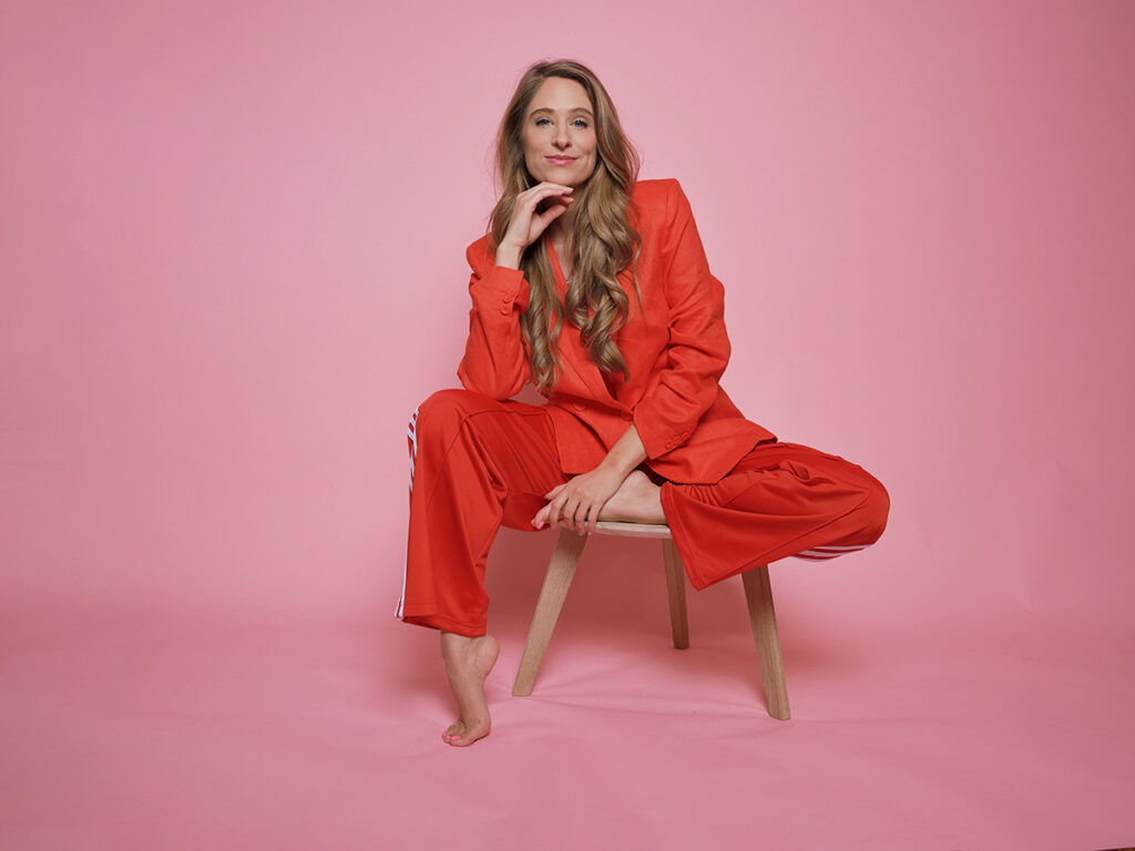 Frau im orangen Anzug auf einem Hocker vor rosa Hintergrund, entspannte und moderne Portraitaufnahme