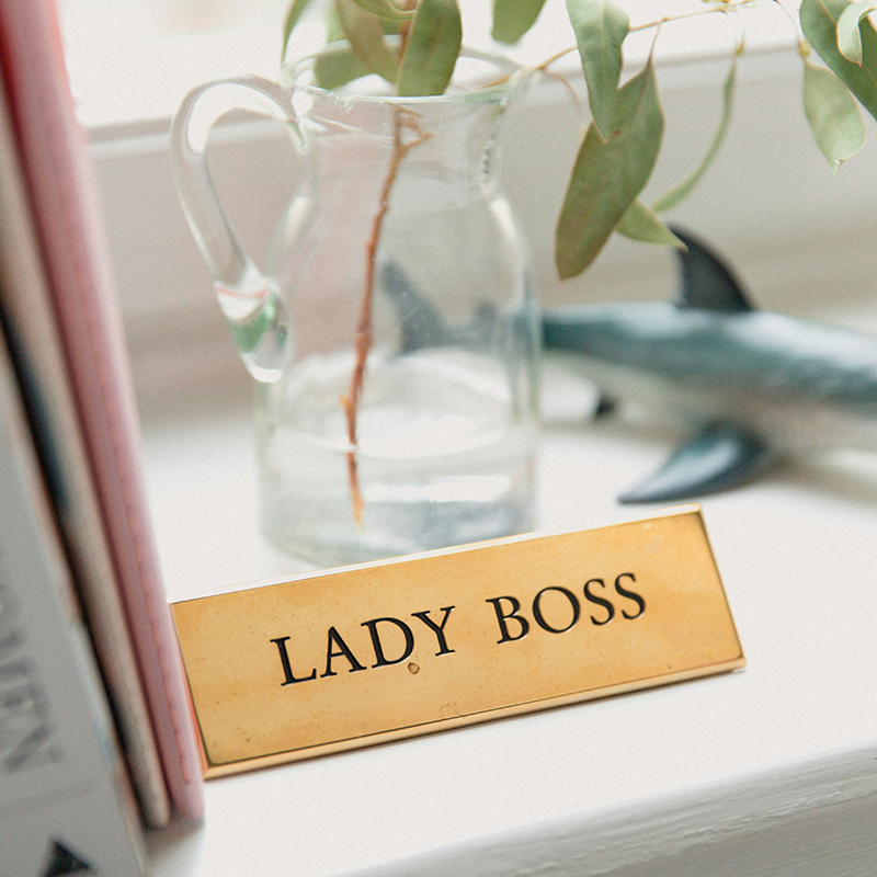 Goldenes Schild mit Aufschrift 'Lady Boss' auf Fensterbank neben Glasvase mit Eukalyptus, inspirierendes Business-Detail.